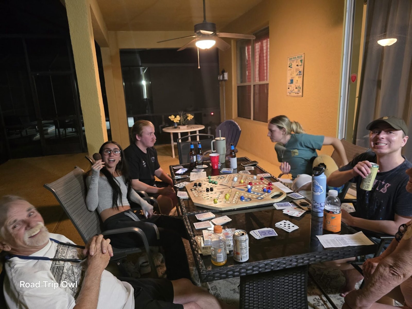 Group of friends and family playing Fast Track marble board game on a patio at night with drinks and cards on the table