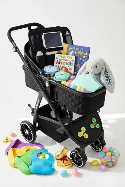Baby road trip Easter basket made from a black 2-in-1 lay-flat stroller filled with a car backseat organizer, spill-proof snack containers, silicone bibs, suction cup spinner toys, Touch and Feel Adorable Animals board book, A Bedtime Full of Stories book, folding potty seat, personalized stuffed bunny, gold foil chocolate Easter bunny, and pastel Easter egg