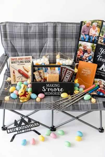 Grandparent camping Easter basket featuring a s'mores caddy with marshmallows and chocolate on a double camping loveseat chair, surrounded by a whittling book, travel bucket list journal, photo blanket, globe string lights, roasting forks, personalized campsite sign, chocolate Easter bunny, and pastel Easter eggs