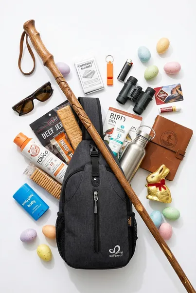 Grandparent hiking Easter basket featuring a crossbody sling bag with a walking stick, beef jerky, peanut butter crackers, Grand Tongo bug spray, anti-chafe balm, emergency blanket, whistle, binoculars, bird guide, hiking journal, energy bars, water bottle, sunglasses, National Parks pass, chocolate Easter bunny, and pastel Easter eggs