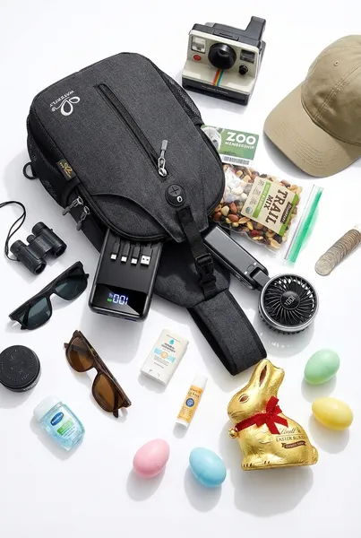 Grandparent zoo Easter basket made from a crossbody sling bag with a portable charger, Polaroid camera, handheld fan, binoculars, trail mix, baseball cap, zoo membership card, sunglasses, sunscreen, hand sanitizer, chocolate Easter bunny, and pastel Easter eggs
