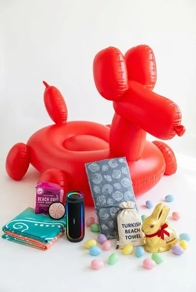 Teen beach Easter basket made from a red balloon dog pool float filled with an oversized sea turtle beach blanket, portable Bluetooth speaker, Beach Day coconut scented sand remover, Barine Turkish beach towel, gold foil chocolate Easter bunny, and pastel Easter eggs