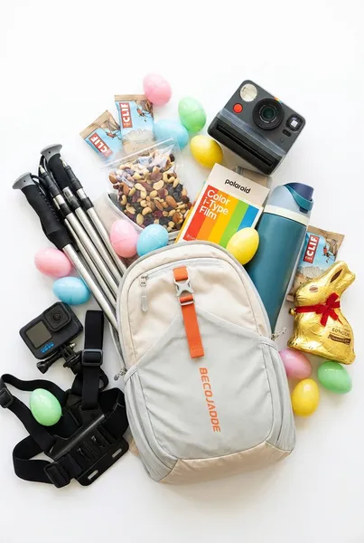 Teen hiking Easter basket made from a beige Becojadde foldable hiking backpack filled with trekking poles, GoPro action camera, Polaroid instant camera, Owala water bottle, trail mix, Clif bars, gold foil chocolate Easter bunny, and pastel Easter eggs