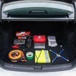 A neatly organized car emergency kit laid out in an open trunk including a jump box, tire inflator, road flares, first aid kit and gloves