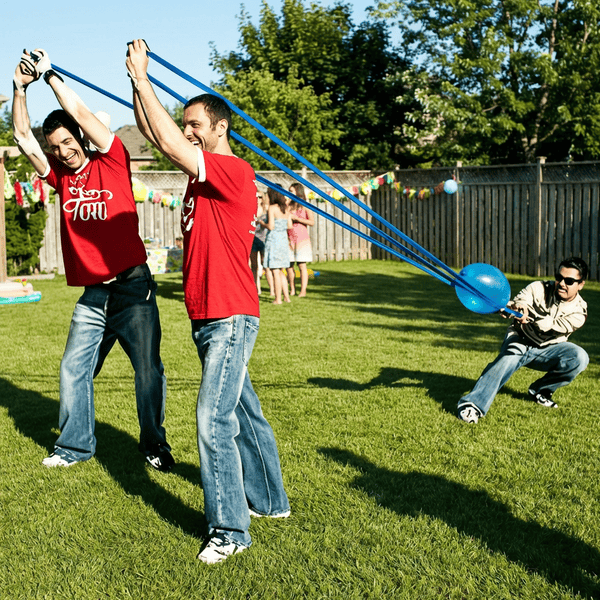 Three adults stretching a giant water balloon slingshot launcher in a sunny backyard