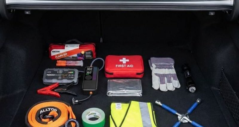A neatly organized car emergency kit laid out in an open trunk including a jump box, tire inflator, road flares, first aid kit and gloves