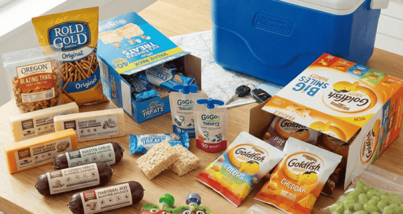 Ultimate road trip snack spread on a kitchen table including pretzels, cheese, summer sausage, Goldfish crackers, Rice Krispies Treats, GoGo Squeez yogurt pouches, vacuum sealed fresh fruit, and more arranged next to a blue Coleman cooler
