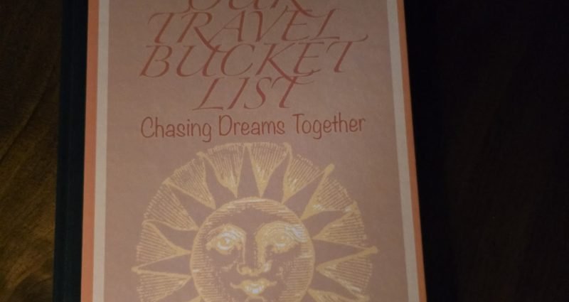 Our Travel Bucket List journal with sun design on hardcover, sitting on wooden table